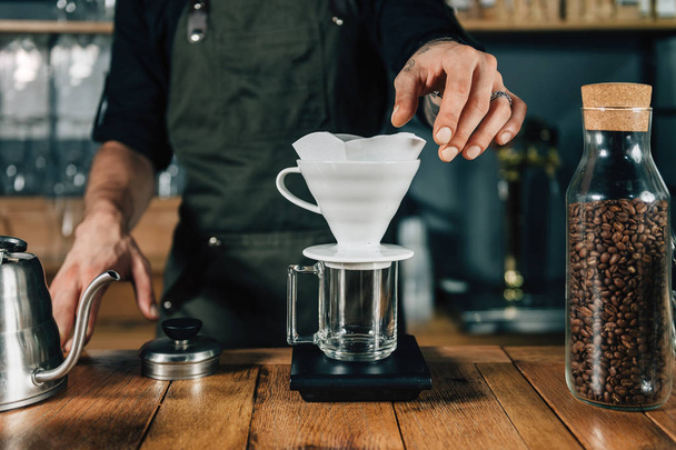 Male barista hands putting white filter to ceramic dripper. Tools and equipment for making Drip Brew coffee on wooden table.  Barista with tattooed arms wearing dark uniform.    - Foto, afbeelding