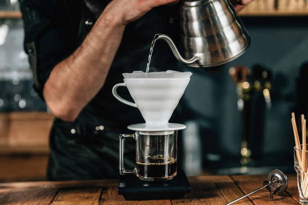 Nahaufnahme eines jungen männlichen Baristas, der kochendes Wasser aus einem Wasserkocher in eine tropfende Kaffeemaschine auf einem Holztisch gießt. Barista in dunkler Uniform.  - Foto, Bild