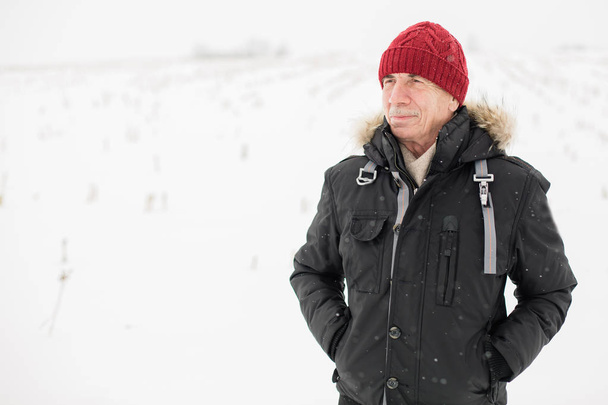 Close up portrait of  senior man standing outdoors in red hat and black jacket winter season. Snowy weather.  - Foto, Bild