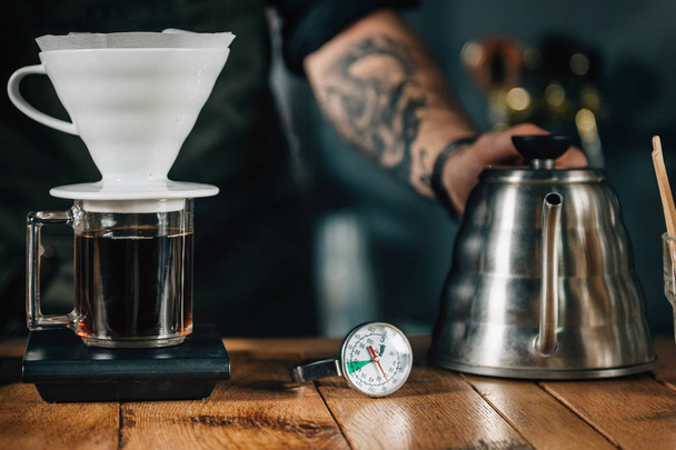 Close up of Drip coffee maker, kettle with thermometer and digital scale on wooden table. Tools and equipment for making Drip Brew coffee. Barista with tattooed arms wearing dark uniform.     - Photo, Image