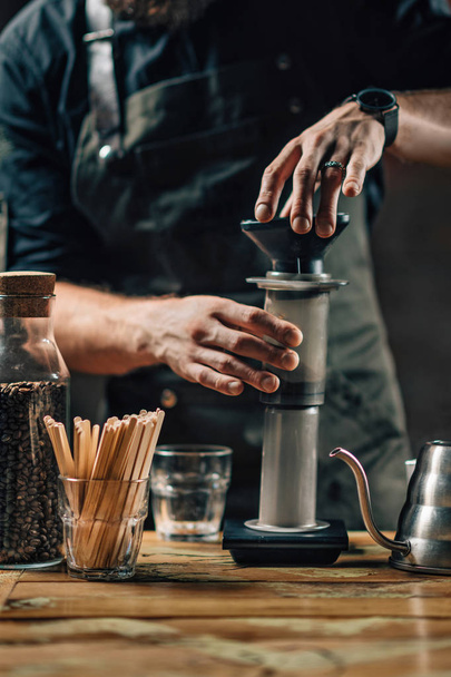 vertikales Bild eines Baristas, der Kaffee aus der Luft zubereitet. Barista mit tätowierten Armen in dunkler Uniform. Holztisch. - Foto, Bild