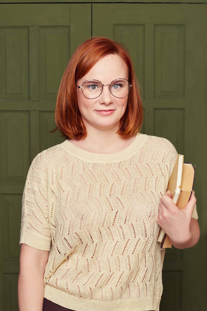 Good looking ginger haired woman with satisfied expression, smiles gently, has intrigued look, thinks about creative plan, studies at college or university, holds books, posing over classroom door. - Zdjęcie, obraz