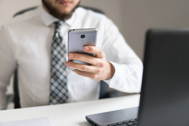 Holding smartphone in hand. Businessman work with computer on table in office work. Young bearded businessman. Office work with a laptop. Financial business. Light background. - Φωτογραφία, εικόνα