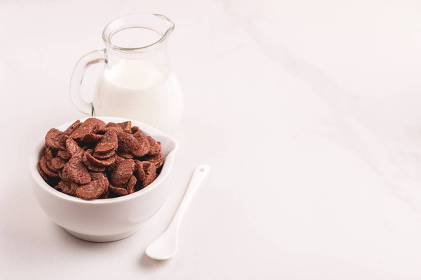 Crunchy chocolate corn flakes in clay bowl with jar of fresh milk on light marble background. Healthy cereals breakfast. Copy space - Foto, Imagem