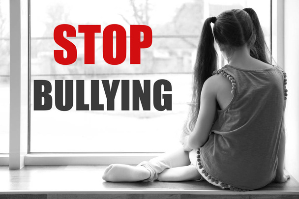 Lonely little girl sitting near window indoors and message STOP BULLYING on background, black-white effect - Foto, imagen