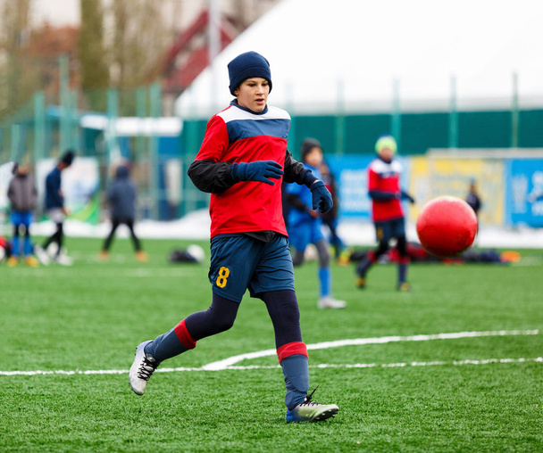 Молоді хлопчики активного спорту в червоно-синьому спортивному одязі бігають і штовхають червоний м'яч на футбольне поле зі штучним дерном. Футбольна молодіжна команда грає у футбол. заняття для дітей, навчання
 - Фото, зображення