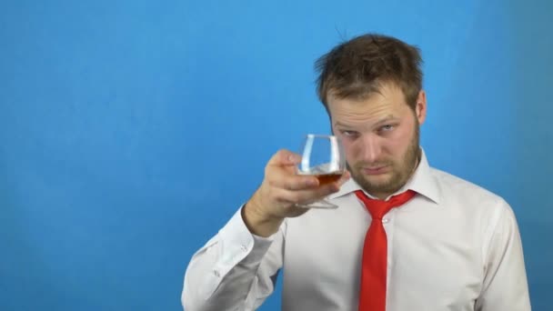 Drunk caucasian man with a beard in a white shirt and tie drinking alcohol brandy from a glass, blue background, hangover - Footage, Video
