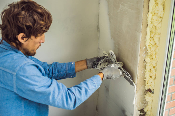 Young man smearing putty on wall while renovating house in daytime. - Fotoğraf, Görsel