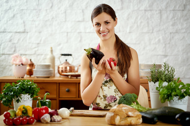 όμορφη νεαρή γυναίκα μελαχρινή κατέχει μελιτζάνα και ντομάτα στην κουζίνα σε ένα τραπέζι γεμάτο από βιολογικά λαχανικά - Φωτογραφία, εικόνα