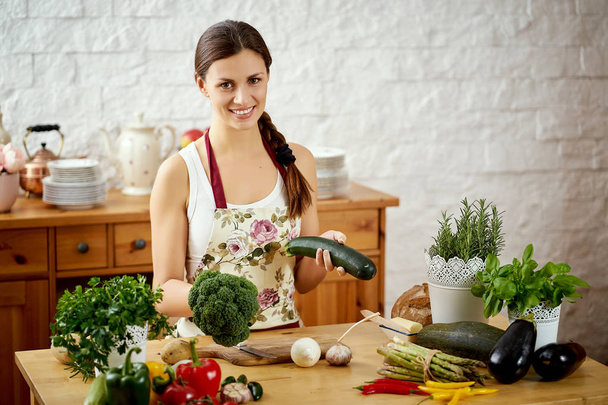 молода брюнетка, що тримає кабачки та брокколі на кухні за столом, повним органічних овочів
 - Фото, зображення