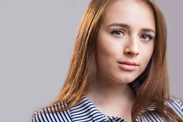 Close up face of a beautiful young girl with a clean skin. A young woman with beautiful eyebrows, long eyelashes, plump lips and long red hair looks at the camera on a gray background. Natural beauty - Foto, Imagen