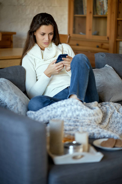 beautiful young smiling woman in white sweater texting on her phone on a gray sofa - Photo, Image