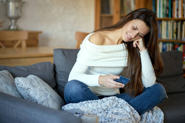 beautiful young smiling woman in white sweater texting on her phone on a gray sofa - Foto, imagen