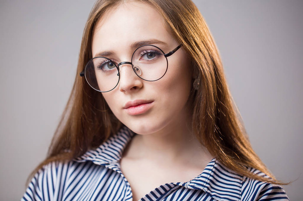Beautiful closeup portrait of a young student girl wearing glasses on a gray background. Attractive redhead woman with natural beauty looks at the camera and smiles. Youth, clean skin, facial care - Fotoğraf, Görsel