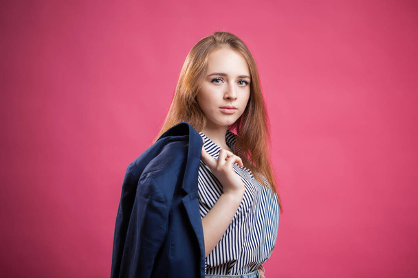 Fashionable young woman posing on a pink background with copy space. Student girl holding his jacket behind his back and looking at the camera - Photo, Image