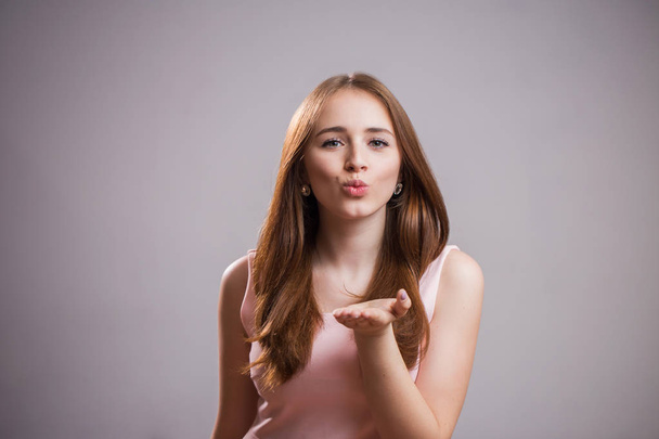 Beautiful young woman wears a pink dress and sends a kiss to the camera on a gray background with copy space for text - Photo, Image