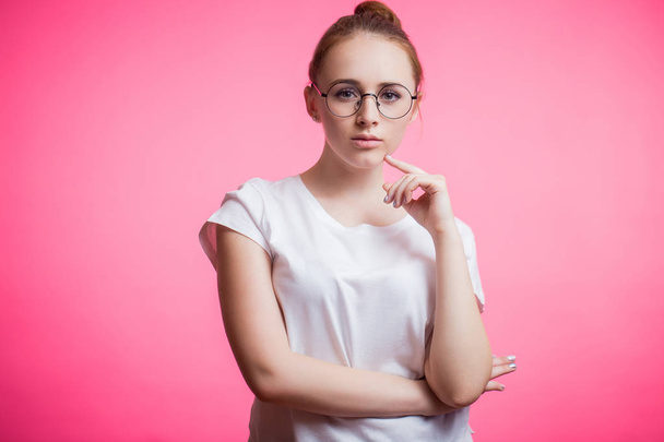 Horizontal portrait of a beautiful young woman wearing round glasses, a white shirt and looking at camera on a pink background with copy space - Fotografie, Obrázek