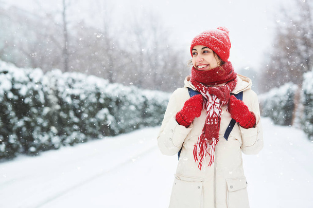 Cheerful smiling woman in warm clothes, red knitted cap, scarf and mittens walking on the snowy street under falling snowflakes after blizzard in city. Happy woman playing with the snow - Photo, image