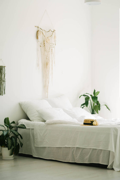Lichte kamer. Interieur design. Groot bed met een deken. Kussens. Comfort, eenvoud. Groot raam en planten. Rieten hoed. - Foto, afbeelding