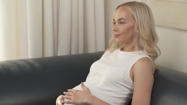 A side view of a lovely pregnant blonde sitting on the sofa and stroking the belly. She sighs, smiles slightly looking at it. She looks in front of her happily - Imágenes, Vídeo