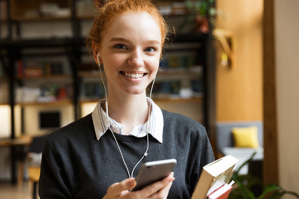 Image of beautiful cheerful happy redhead lady student posing indoors in library holding books listening music with earphones using mobile phone. - Photo, Image
