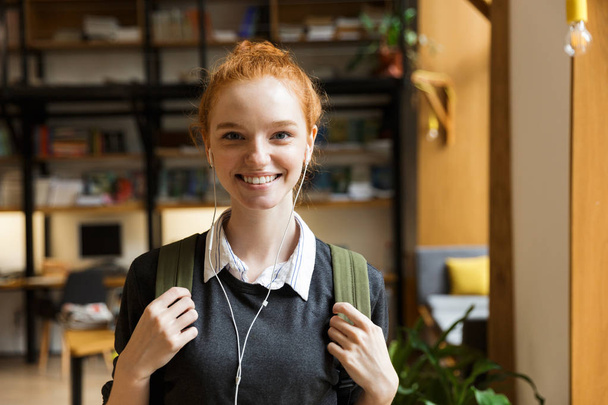 Bild der schönen rothaarigen Studentin, die drinnen in der Bibliothek posiert und Musik mit Kopfhörern anstimmt. - Foto, Bild