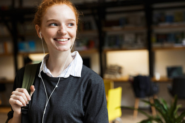 Smiling younh teenage girl in earphones carrying backpack in library - Foto, Imagen