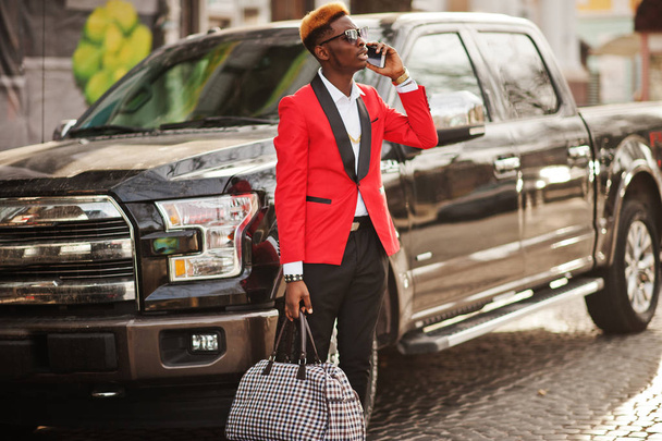 Fashion african american man model at red suit, with highlights hair posed against big suv black car and speaking on mobile phone. - Fotó, kép