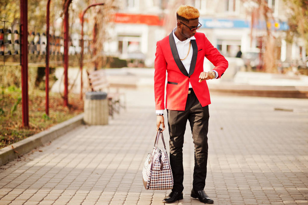 Fashion african american man model at red suit, with highlights hair and handbag posed at street and looking at his watches. - Фото, изображение
