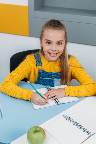 fröhliches Kind schreibt während des Unterrichts mit Apfel auf Schreibtisch in Notizblock  - Foto, Bild