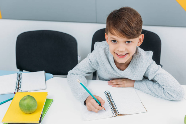 happy schoolboy writing in notepad in classroom - Фото, изображение