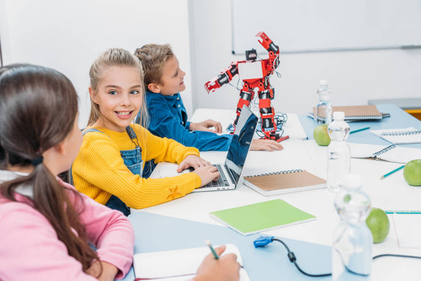 niños escribiendo en el cuaderno y usando el ordenador portátil en el escritorio con robot rojo en la clase del tallo
 - Foto, imagen