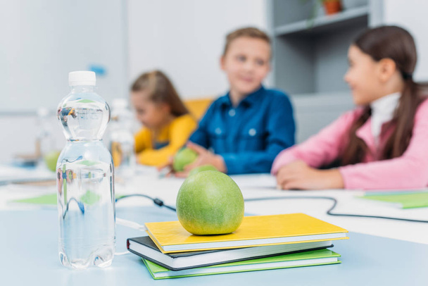 green ripe apple on notebooks and schoolchildren at background in classroom - Photo, Image