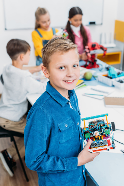 smiling schoolboy holding robot and looking at camera with classmates at background in classroom - Foto, immagini