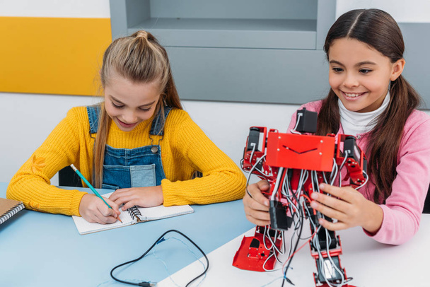 Улыбающиеся школьницы, пишущие в блокноте и держащие красного робота за столом в классе по образованию
 - Фото, изображение