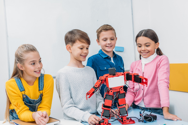 smiling schoolchildren looking at red robot handmade on desk in stem class - Фото, изображение