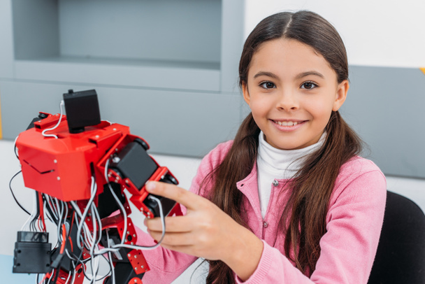 close up view of schoolgirl smiling, holding red handmade robot and looking at camera in stem class - Foto, Bild
