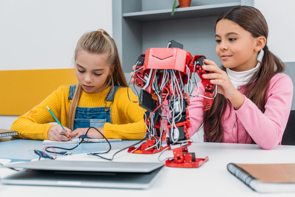 schoolgirls writing at notebook and touching red electric robot during stem lesson - Valokuva, kuva