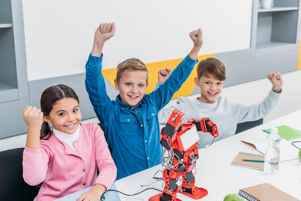 Kinder jubeln und heben während der Vorlesung mit Elektroroboter die Hände am Schreibtisch - Foto, Bild