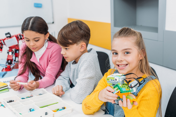 улыбающаяся школьница с разноцветным роботом в руках, в то время как одноклассники смотрят на коробку с деталями
 - Фото, изображение