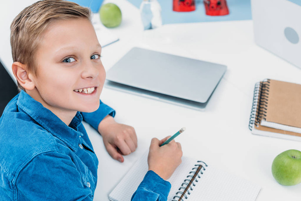 smiling boy writing in notebook, smiling and looking at camera in stem class - Foto, Imagen