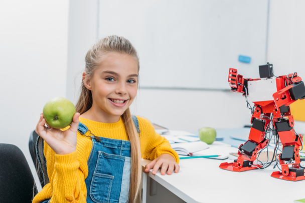 hymyilevä koulutyttö istuu pöydässä robottimallin kanssa STEM luokkahuoneessa, pitäen omenaa ja katsoen kameraa
 - Valokuva, kuva