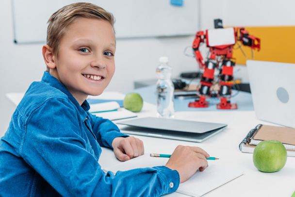 schoolboy sitting at desk with robot model, looking at camera and writing in notebook during STEM lesson - Photo, Image