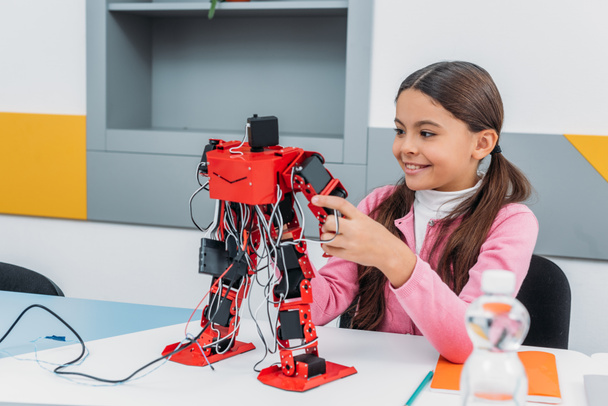 adorable schoolgirl sitting at table, smiling and holding robot model during STEM lesson   - Foto, afbeelding