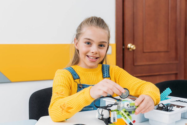 жизнерадостная школьница смотрит в камеру и работает над моделью робота ручной работы во время урока STEM
   - Фото, изображение