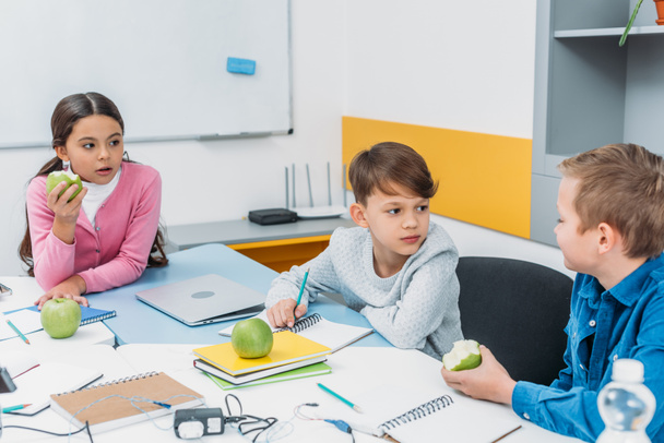 Mitschüler sitzen am Tisch, machen Pause, reden und essen Äpfel im Klassenzimmer - Foto, Bild