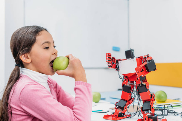 школярка їсть яблуко і сидить за столом з моделлю робота в класі STEM
 - Фото, зображення