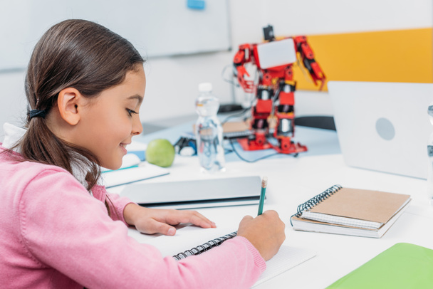 schoolgirl sitting at desk with robot model and writing in notebook during STEM lesson - Foto, Imagem