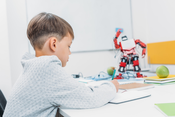 schoolboy sitting at desk with robot model and writing in notebook during STEM lesson - Foto, Imagen