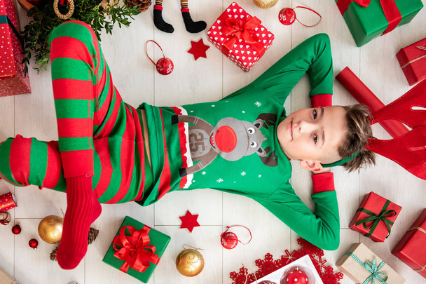 Cute young boy wearing xmas pajamas lying on the floor, surrounded by christmas presents, ornaments and decorations, top view. - Fotoğraf, Görsel